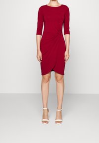 Frau trägt ein knielanges rotes Kleid mit Dreiviertelärmeln und seitlicher Knoten-Detail, kombiniert mit weißen High Heels Sandalen.