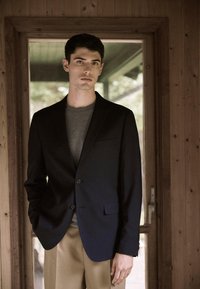 Laevastiku sinine pintsak notiga kraega ja kahe nööbiga, kombineeritud halli klassikalise kaelusega särgi ja beežide pükstega. Taustal on puit.