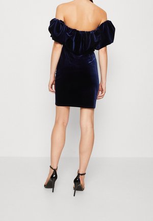 Kobieta stojąca tyłem, ubrana w granatową aksamitną sukienkę z odkrytymi ramionami oraz czarne szpilki z paskiem wokół kostki.