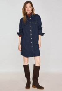 Robe chemise en denim bleu foncé, avec un devant à boutons, des poches poitrine, des manches retroussées et un ourlet arrondi, associée à des bottes marron montantes.