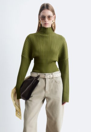 Femme portant un pull à col roulé côtelé vert olive, pantalon beige avec ceinture, tenant un sac noir et une écharpe à motifs, avec lunettes de soleil et boucles d'oreilles.