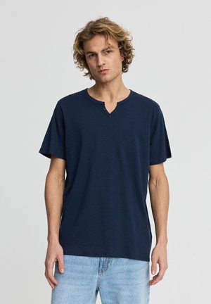 Giovane uomo con capelli biondi ricci che indossa una maglietta henley blu navy a maniche corte e jeans azzurri, in piedi davanti a uno sfondo semplice.