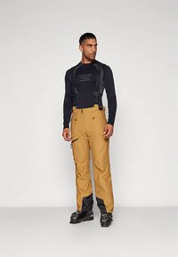 Brune, vanntette skibukser med svarte detaljer nederst, justerbare stropper, flere lommer og en tilpasset silhuett. Brukt sammen med en svart langermet skjorte.