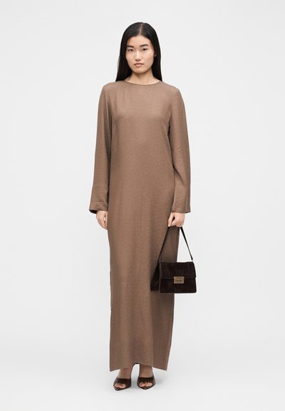 Γυναίκα που φορά ένα μακρυμάνικο, καφέ φόρεμα μέχρι τον αστράγαλο, κρατώντας μια σκουρόχρωμη καφέ τσάντα και φορώντας ανοιχτά καφέ παπούτσια, στέκεται πάνω σε λευκό φόντο.