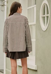 Camicia a righe marrone e crema con bottoni, taglio oversize con maniche lunghe e dettagli ai polsini, abbinata a una gonna nera plissettata.