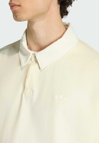 Jasnokremowa koszulka polo z kołnierzykiem, z subtelnym haftowanym logo, gładką fakturą i detalami z guzikami.