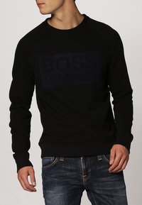 Sudadera negra con cuello redondo, que presenta una impresión oscura de "BOSS ORANGE" en el frente. Mangas y dobladillo acanalados. Combinada con jeans azules.