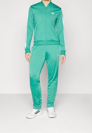 Personne portant un survêtement vert à fermeture éclair intégrale avec des bandes blanches sur les manches et les jambes, assorti de baskets blanches à rayures noires.