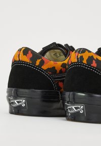 Svarta mockaskor med ett överdel i orange och svart leopardmönstrad kanvas. Har vit sömnad och en svart gummisula med en logotyp.