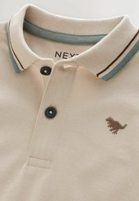 Polo beige avec un tissage texturé, ornée d'un col rayé bleu clair et marron, de deux boutons noirs et d'une broderie de dinosaure marron.