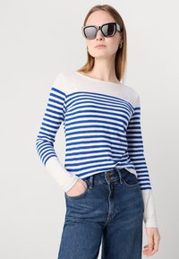 Kék és fehér csíkos hosszú ujjú póló, hajónyakú kivágással, magas derekú farmernadrággal párosítva. A modellen oversized napszemüveg van.