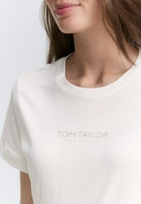Bílý bavlněný tričko s kulatým výstřihem, které na přední straně zdobí jemný šedý potisk "TOM TAILOR EST - 1962". Měkká, hladká textura.