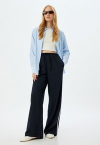  Lichtblauw button-up shirt over een witte cropped top, gecombineerd met marineblauwe wijde broek met witte zijstrepen en witte sneakers.