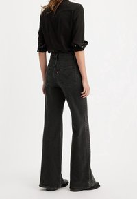 Jeans noirs évasés taille haute avec une finition texturée, dotés de poches arrière et d'une étiquette de logo rouge, portés avec une chemise noire à boutons.