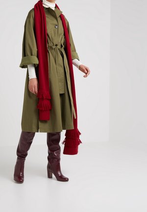 Personne portant un trench-coat ceinturé vert olive, un col roulé blanc, une longue écharpe rouge avec des franges et des bottes à talons bordeaux montant jusqu'aux genoux.