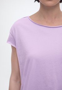 Γυναίκα που φοράει ανοιχτό μωβ κοντομάνικη μπλούζα με στρογγυλή λαιμόκοψη, εμφανιζόμενη από τους ώμους έως το μέσο του κορμού με φόντο λευκό.