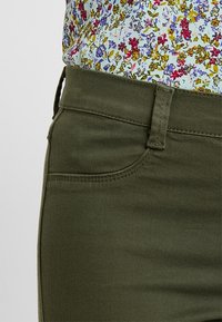 Olivgrüne Denim-Jeans mit Slim Fit, vordertasche und subtilen Stickdetails, kombiniert mit einem hellen, Blumenmuster-Hemd.