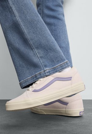 Personne portant des baskets violet clair avec une bande lavande et un jean bleu, debout sur un sol gris contre un fond clair.