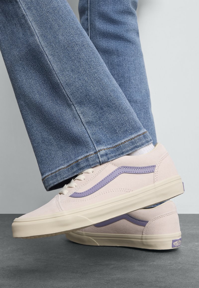 Personne portant des baskets violet clair avec une bande lavande et un jean bleu, debout sur un sol gris contre un fond clair.