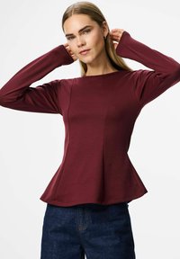 Sieviete burgundijas krāsas garām piedurknēm blūzē ar paplatinātu apakšdaļu, pielāgojot matus, tērpusies tumši zilos džinsos uz vienkārša balta fona.