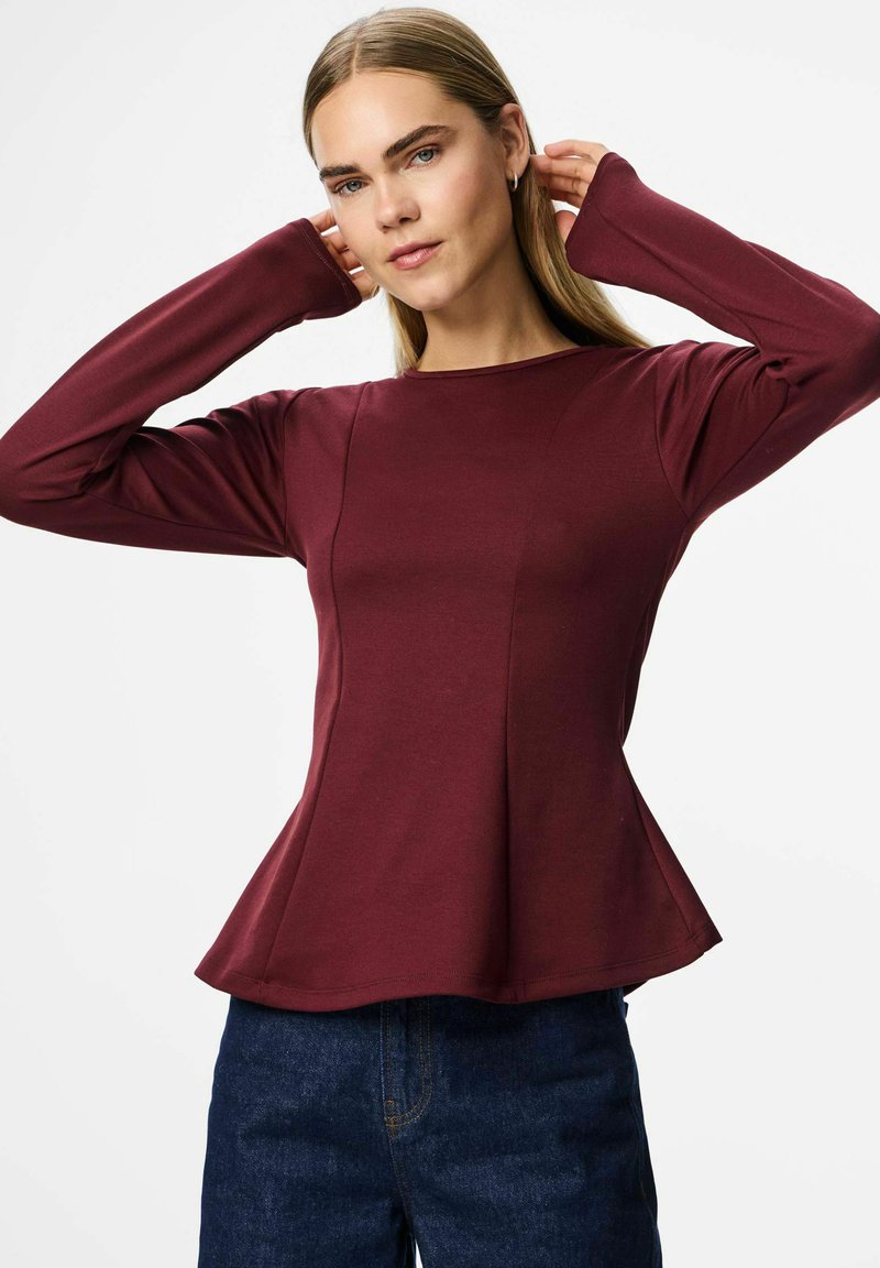 Sieviete burgundijas krāsas garām piedurknēm blūzē ar paplatinātu apakšdaļu, pielāgojot matus, tērpusies tumši zilos džinsos uz vienkārša balta fona.