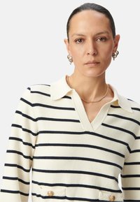 Gestreifter Pullover in Creme mit marineblauen Streifen, ausgestattet mit einem Kragen, V-Ausschnitt und goldenen Knopfdetails an den vorderen Taschen.