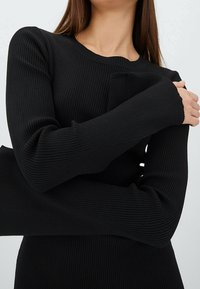 Femme croisant les bras sur la poitrine, portant un haut noir à manches longues côtelé avec des manches prolongées, sur un fond neutre.