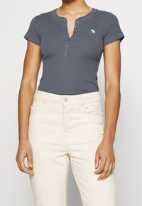 Snäv grå ribbad t-shirt med korta ärmar, tryckknappsstängning och en liten vit logotyp. Kombinerad med ljusbeiga jeans.