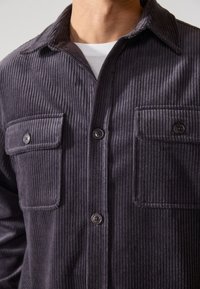 Chemise en velours côtelé gris foncé avec un devant à boutons, deux poches poitrine et un col classique. Présente une texture à côtes verticales et des boutons noirs.