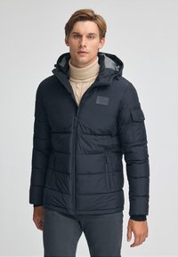 Svart puffjacka med huva, med en strukturerad finish, fickor med dragkedja och ett märke på bröstet. Bärs över en ljus polotröja.