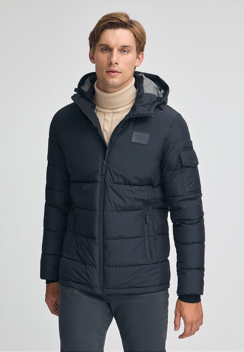 Svart puffjacka med huva, med en strukturerad finish, fickor med dragkedja och ett märke på bröstet. Bärs över en ljus polotröja.