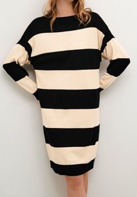 Femme portant une robe en tricot à rayures horizontales larges noir et beige à manches longues, se tenant avec les mains sur les hanches devant un fond uni.