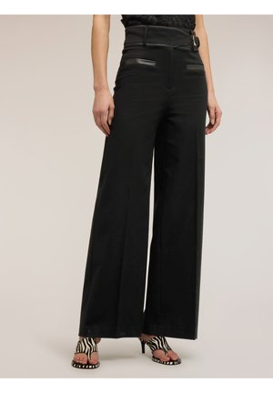 Femme portant un pantalon noir large à taille haute avec ceinture et un haut en dentelle noire, associé à des sandales à talons ouvertes à motif zébré.