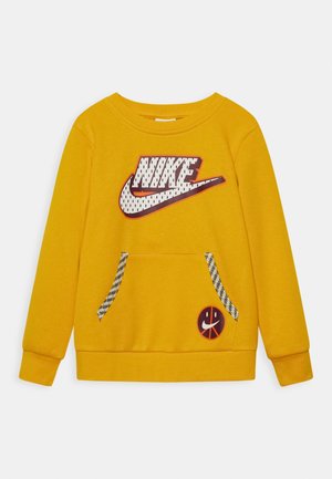Sweat-shirt jaune à manches longues avec un grand logo Nike blanc et marron sur la poitrine, des bords de poche à motif, et un petit patch smiley basket-ball sur la poche avant.