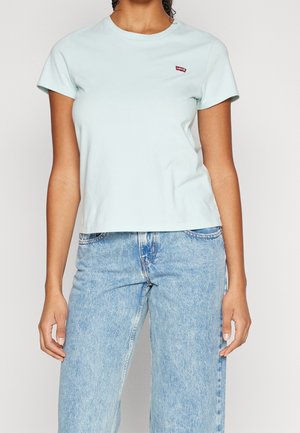 Kobieta ubrana w jasnoniebieską koszulkę Levi's z krótkim rękawem oraz jasne dżinsy z wysokim stanem, stojąca na jednolitym tle.