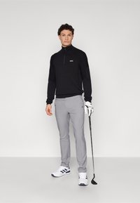 Črno zip-up pulover, sivi golf hlače, bele čevlje z mornarsko modrimi poudarki, drži golf palico, pozira proti enotni ozadju.