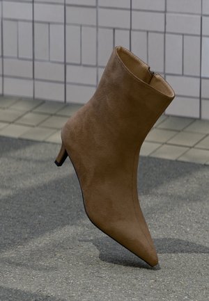 Brun ankelstøvlett i semsket skinn med spiss tå, glidelåslukking og lav stiletthæl, vist mot en teksturert bakgrunn.