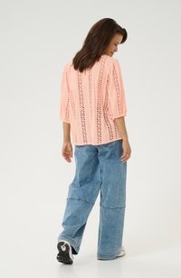 Top rosa con detalles de ojales, mangas de tres cuartos, combinado con jeans de denim sueltos. El conjunto presenta un corte relajado y una tela texturizada.