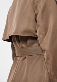 Brun trenchcoat med belte i midjen, strukturerte skuldre og et lagdelt design. Har en glatt, matt tekstur og plisserte detaljer.