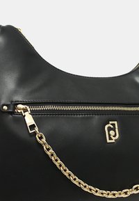 Bolso de mano de cuero negro con cremallera dorada y detalle de cadena, que presenta un emblema de logo dorado en el frente.