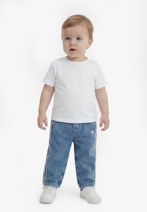 Tout-petit aux cheveux châtain clair portant un t-shirt blanc, un jean de survêtement Adidas bleu et des baskets blanches, debout sur un fond blanc.