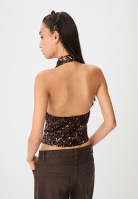 Hnedý halter top s flitrami a dizajnom s otvoreným chrbtom. Top má textúrovaný povrch a je kombinovaný s tmavými nohavicami s vysokým pásom.