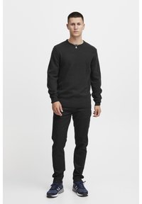 Temno siva dolgosleeve pulover z rebrastimi manšetami in robom, nosi ga z črnimi hlačami in modrimi supergami, na nevtralni ozadju.