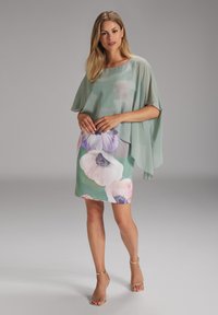 Un vestito floreale verde presenta un overcape trasparente. La gonna ha grandi stampi floreali pastello con accenti circolari e una silhouette aderente.