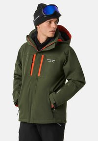 Grüne Softshelljacke mit orangefarbenen Akzenten, mehreren Reißverschlusstaschen, verstellbaren Manschetten und Kapuze. Trägt man über einer schwarzen Basisschicht und Hose.