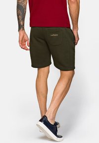 Ciemnooliwkowe szorty z miękkiego materiału, z podwiniętymi nogawkami i tylną kieszenią z logo. Noszone z czarnymi trampkami.