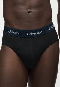 Κοντινή λήψη του κορμού ενός ατόμου με μαύρα εσώρουχα Calvin Klein και μπλε ζώνη μέσης που εμφανίζει το όνομα της μάρκας με λευκά γράμματα.