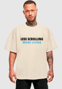 Beige oversize tričko s krátkým rukávem má na přední straně černý a modrý nápis: „MÉNĚ SKROLLINGU, VÍCE ŽIVOTA.“