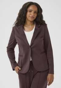 Blazer bordeaux avec un col à revers, fermeture à un bouton, deux poches avant et manches longues, associé à un haut blanc. Texture de tissu lisse.