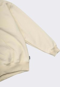 Felpa color crema con tessuto strutturato, dotata di tasca frontale e polsini a coste. Un lato presenta un piccolo etichetta con il logo.